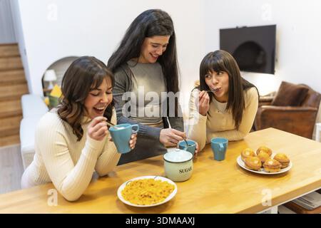 Drei junge Erwachsene Freundinnen genießen Frühstück mit Muffins, Kaffee und Müsli, Lächeln und Lachen an einem Holztisch in einem gemütlichen Zuhause Stockfoto