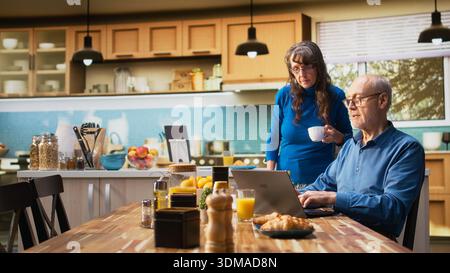 Senioren im Ruhestand genießen morgens die Routine, auf dem Laptop zu surfen, mit hausgemachtem Frühstück auf dem Tisch, bei dem frischer Kaffee und Gebäck serviert werden. Ehemann und Ehefrau unterhalten sich zwanglos. Kamera A.. Stockfoto