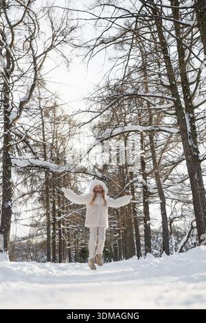Glückliche junge Frau, die auf schneebedeckten Pfaden im Winterwald springt und den kalten, sonnigen Tag draußen genießt. Stockfoto