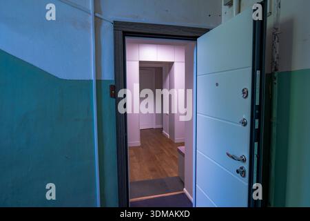 Eine weiße Sicherheitstür steht offen und zeigt warme Holzböden und rosafarbenes Umgebungslicht, von einer abgenutzten, türkisfarbenen und weißen Treppe mit eleganten Befestigungsteilen aus gesehen Stockfoto