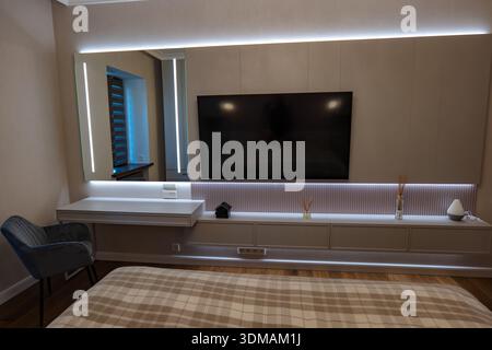 Das moderne Schlafzimmer verfügt über einen an der Wand montierten Flachbildfernseher in beigefarbenem Panel mit LED-Streifen, einen schwebenden Schreibtisch mit beleuchtetem Spiegel und grauen Samtstuhl und Stockfoto