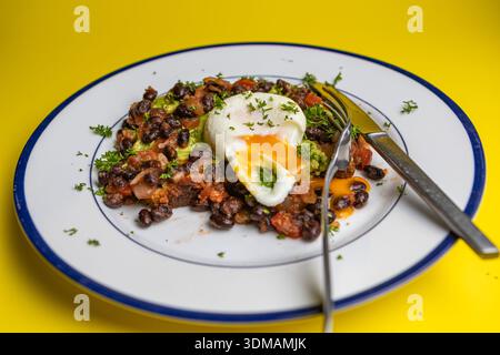 Pochiertes Ei auf Vollkornbrot mit Avocado und schwarzen Bohnen und Tomatensalsa auf einem Teller mit blauem Rand. Auf gelbem Hintergrund Stockfoto