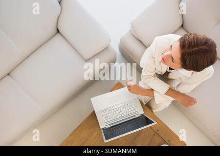 Reife Frau, die Blazer trägt, sitzt auf Sofa zu Hause mit Laptop auf Couchtisch, Kopierraum. Minimalismus, Ruhe, Arbeitsplatz, neutral, Lounge, Fokus, Profi Stockfoto