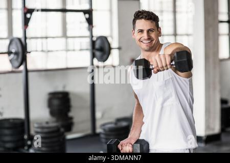Mann in den Zwanzigern, der Tanktop trägt, im Fitnessstudio vorne hochfährt, Hexenhantel hält, Kopierraum. Fitness, Workout, sportlich, Gewichte, Langhantel, Rack, Stockfoto