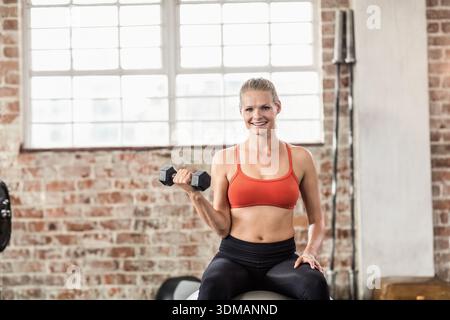 Eine Frau, die auf einem Stabilitätsball sitzt und eine Hexenhantel hält, während sie im Fitnessstudio einen orangefarbenen Sport-BH trägt. Fitness, Kraft, Training, Gewichte, Industrie, Stockfoto