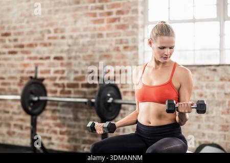 Frau mit orangefarbenem BH, schwarzer Hose, die auf der Bank im Fitnessstudio sitzt und Hexen-Kurzhanteln hält, Kopierraum. Fitness, Training, Kraft, Gewichte, Langhantel, Sonnenlicht Stockfoto