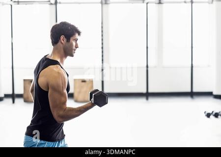 Mann mit schwarzem ärmellosem Top, der einarmige Kurzhantelcurl im Fitnessstudio mit Hexenhantel durchführt. Training, Fitness, Kraft, Tageslicht, Studio, minimal Stockfoto