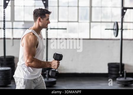 Mann Ende 20, der im Fitnessstudio stand und eine Hexenhantel am Hockgestell hielt, Tank trug, Kopierraum. Industrie, Kraft, Fitness, Training, Sportler, barbell, Stockfoto