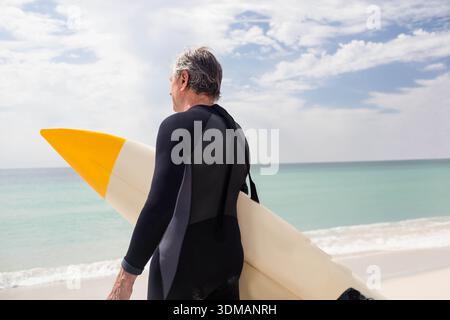 Senior Mann, der am weißen Sandstrand steht und Surfbrett mit gelber Spitze hält und einen Neoprenanzug trägt. Älterer, Board, Longboard, Ocean, Türkis, ruhig, Schauer Stockfoto