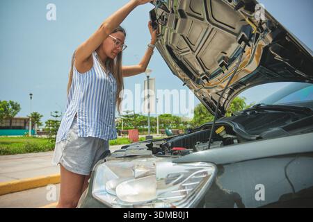 Frau, die neben ihrem kaputten Auto steht, mit geöffneter Motorhaube, und den Motor inspiziert Stockfoto