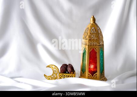 Eine hohe goldene Laterne mit bunten Glas steht neben Datteln auf weißem Seidenhintergrund. Stockfoto