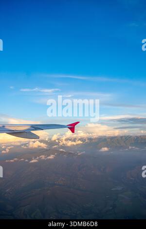 Der Blick aus einem Flugzeug fängt einen Flügel mit einer roten Spitze über sanften grünen Bergen und weichen Wolken unter einem leuchtend blauen Himmel ein. Stockfoto