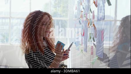Die rothaarige Frau in gestreiftem Oberteil hält blaue Haftnotiz und Stift und ordnet Notizen auf Glasbrett an Stockfoto