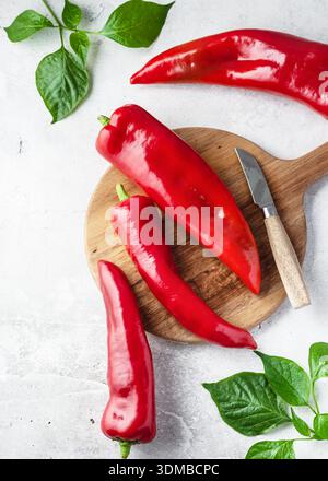 Frische rote Kapia-Paprika (Capsicum annuum) auf einem Holzschneidebrett mit einem Messer, umgeben von grünen Blättern. Stockfoto
