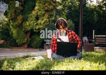 Der Laptop liegt auf den Beinen. Der Student ist im öffentlichen Park draußen. Stockfoto