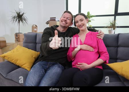 Fröhliches Paar feiert den Meilenstein der Schwangerschaft und entspannt sich auf einer gemütlichen Couch in ihrem modernen Wohnzimmer mit Pflanzen und warmen Farben Stockfoto