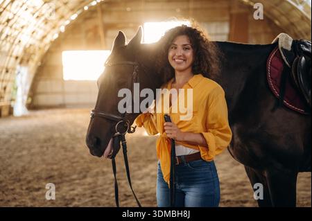 Stehen und posieren. Die schöne junge Frau ist mit einem Pferd im Hangar. Stockfoto