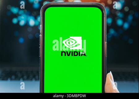 Smartphone mit grünem Bildschirm, auf dem das NVIDIA-Logo angezeigt wird, in der Hand gehalten mit verschwommenem Laptop und Bokeh-Lichtern im Hintergrund. Konzept der künstlichen in Stockfoto