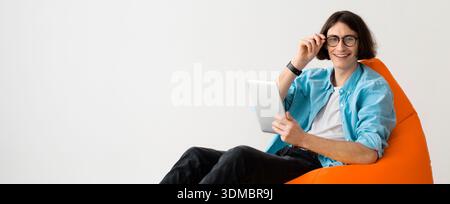 Junger Mann, der auf einem Sitzsack sitzt, während er das Tablet in einem einfachen Innenraum benutzt Stockfoto