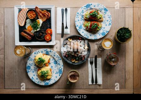 Blick von oben auf einen stilvollen Brunch-Tisch mit englischen Frühstücksprogalen, benedict-Waffeln, Avocado, Toast und Kaffeespezialitäten auf einem Wald Stockfoto