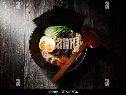 Eine Schüssel Ramen mit einem Löffel und Eiern. Der Ramen wird mit Eiern und Gemüse belegt, und der Löffel ist bereit, um die köstliche Mahlzeit zu genießen Stockfoto
