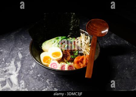 Eine Schüssel Ramen mit einem Löffel drin. Die Schüssel ist mit Nudeln, Fleisch und Gemüse gefüllt Stockfoto