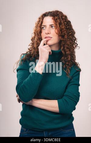 Studio-Porträt einer jungen rothaarigen Frau in grünem Rollkragen und Jeans. Sie schaut nachdenklich auf, ihren Finger auf ihren Lippen, gegen eine Kastration Stockfoto