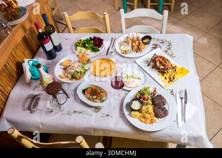 Eine reichhaltige Auswahl an authentischen griechischen und mediterranen Gerichten, einschließlich gegrilltem Fleisch, Meeresfrüchten und Salaten, auf einem wunderschön gedeckten Tisch in einem ungezwungenen Ambiente Stockfoto