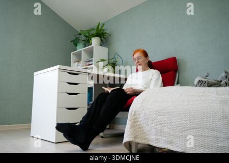 Eine 70-jährige Frau sitzt bequem in einem Sessel zu Hause, tief vertieft in das Lesen eines Buches. Das gemütliche Ambiente spiegelt ein friedliches Hobby im Ruhestand wider Stockfoto