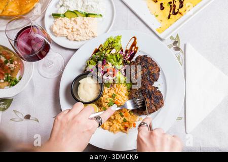 Ein lebhafter Top-Down-Schuss fängt eine Person ein, die Fleisch mit Besteck auf einem Teller mit mediterranen Fleischbällchen und Reis schneidet, während ihre Hand auch ein Glas hält Stockfoto