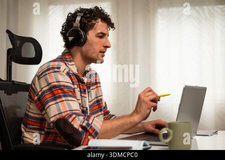 Moderner junger Mann, der kabellose Kopfhörer und Laptop nutzt, während er bequem von zu Hause aus an E-Learning-Sitzungen teilnimmt. Stockfoto