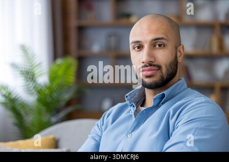 Selbstbewusster junger erwachsener Mann mit Bart und hellem Hemd sitzt entspannt in einem gemütlichen Heimbüro und repräsentiert professionelle Lifestyle- und Work-from-Home-Konzepte Stockfoto