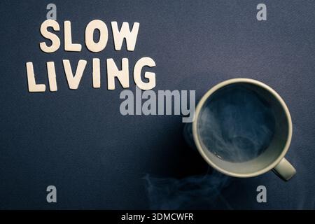 Slow Living Konzept mit einer dampfenden Kaffeetasse und Holzbuchstaben, die Slow Living auf dunklem Hintergrund beschreiben. Minimales Stillleben, das den Geist repräsentiert Stockfoto