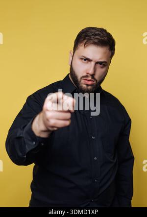 Ein Mann steht in einem dunklen Hemd und zeigt direkt auf die Kamera. Er hat einen fokussierten Ausdruck im Gesicht. Der Hintergrund ist hellgelb, Additiv Stockfoto