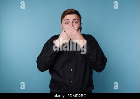 Ein Mann mit kurzen braunen Haaren steht vor blauem Hintergrund. Er bedeckt seinen Mund mit beiden Händen und zeigt einen Ausdruck der Überraschung. Sein schwarzes Hemd Stockfoto