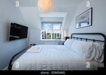 Ein Schlafzimmer mit einem weißen Bett und einem schwarz gerahmten Bild an der Wand. Das Bett ist ordentlich gemacht und es gibt einen Fernseher an der Wand. Das Zimmer hat Stockfoto