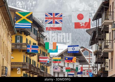 Internationale Flaggen hängen über einer verschneiten Straße in Cortina d Ampezzo, die vor alpinen Chalets und nebeligen Bergen steht und die globale Einheit, den Winter, symbolisiert Stockfoto
