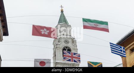 Internationale Flaggen hängen über einer verschneiten Straße in Cortina d Ampezzo, die vor alpinen Chalets und nebeligen Bergen steht und die globale Einheit, den Winter, symbolisiert Stockfoto