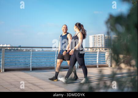 Schwangere Frau und Mann spazieren lässig entlang der Uferpromenade, teilen Lächeln und lachen, genießen eine gute Zeit, während sie einen gesunden Lebensstil genießen Stockfoto