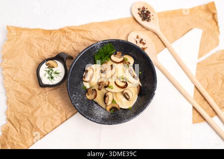 Blick von oben auf mit Pilzen gefüllte Knödel, serviert in einer schwarzen Schüssel mit Sauerrahm, frischen Kräutern und Holzlöffeln auf Pergamentpapier Hintergrund. Stockfoto