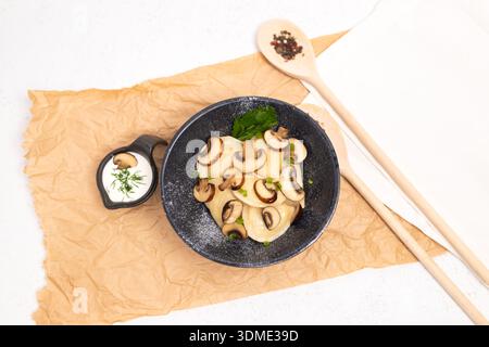 Hausgemachte Teigtaschen mit Pilzfüllung, serviert in einer schwarzen Schüssel mit Sauerrahm und Kräutern. Flach liegende Essenszusammensetzung auf Pergamentpapier. Stockfoto