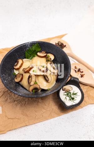 Seitenansicht von Knödeln mit Pilzfüllung, serviert in einer schwarzen Schüssel mit Sauerrahm und frischen Kräutern auf rustikalem Pergamentpapier. Stockfoto