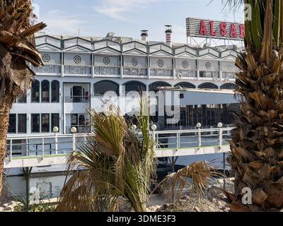 Blick auf ein schwimmendes Restaurant und Architektur am Fluss entlang der Nilküste in Kairo, Ägypten, eingerahmt von Palmen und städtischen Gebäuden am Wasser Stockfoto