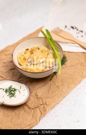 Traditioneller ukrainischer Varenyky, serviert in einer Keramikschale mit gebratenen Zwiebeln, grüner Zwiebelgarnitur und Sauerrahm. Rustikales Essen auf Pergamentpapier, Stockfoto