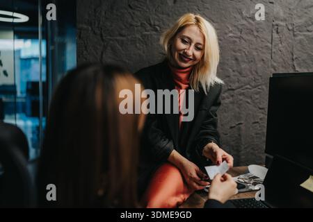 Selbstbewusste Geschäftsfrau in einem schwarzen Blazer lächelt, während sie mit einem Kollegen im Büro über Papiere spricht Stockfoto