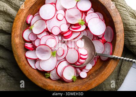 Nahaufnahme von Radieschen in Holzschüssel mit Löffel auf textilem Hintergrund. Frische Salatzubereitung für gesunden Lebensstil und Hausmannskost Stockfoto