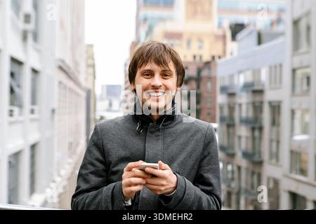 Lächelnder junger Mann, der draußen in einer modernen Stadt ein Smartphone benutzt. Porträt des urbanen Lifestyles mit Mobiltechnologie, Kommunikation, Selbstvertrauen und lässiger Atmosphäre Stockfoto