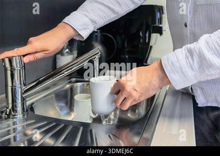 Everyday Utility Woman füllt weißen Becher mit Wasser am Edelstahlwaschbecken, Hände unter fließendem Wasser, Büro-Küchenzeile mit Tassen und Theke Stockfoto
