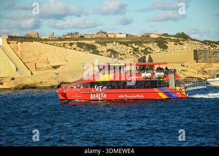Sliema, Malta - 29. November 2025: Ein roter Touristenkatamaran I See Malta segelt entlang der felsigen Küste und alten Steinbefestigungen unter blauem Himmel. Stockfoto