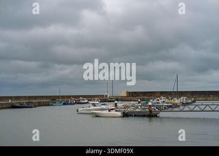 Mehrere Boote liegen an einem dunkelgrau bewölkten Nachmittag im schottischen Hopeman Harbour vor. Eine lange Steinmauer bildet einen Teil des Hafens mit einem Metallpfeiler Stockfoto
